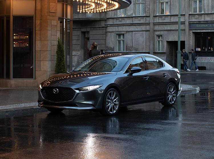 Blue Mazda3 with headlights on parked in front of hotel on dark, wet urban street.