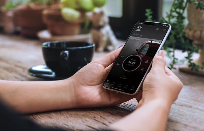 Woman checks her smartphone displaying MyMazda app Remote Start screen at a café table.