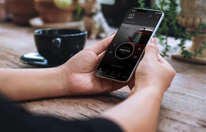 Une femme dans un café consulte son téléphone qui affiche l’écran de démarrage à distance de l’appli MyMazda.