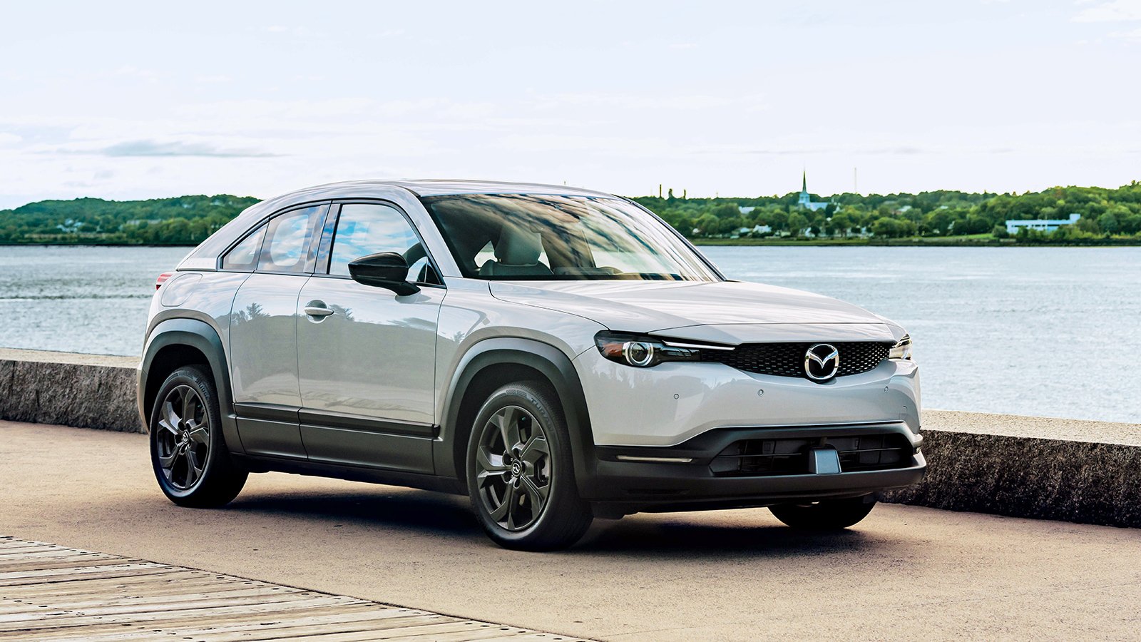 Sunlight and blue cloudy sky reflects off of white MX-30 EV parked by waterfront pier. 