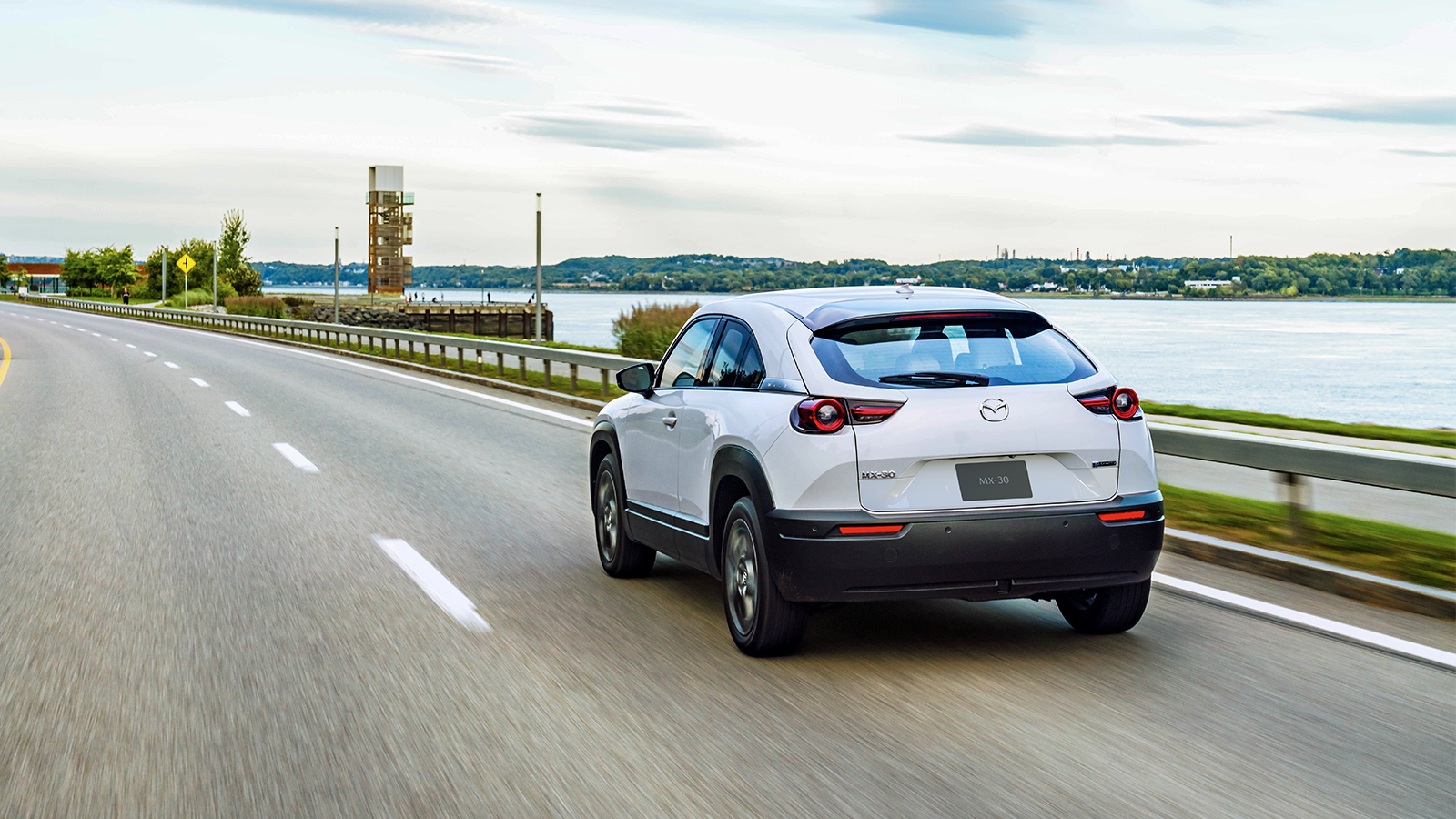 White MX-30 EV drives past blurred waterfront road.