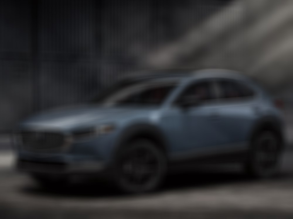 Driver’s side profile of CX-30 in hangar bay, rays of sunlight beam atop the vehicle, dark stacked storage containers line the background. 