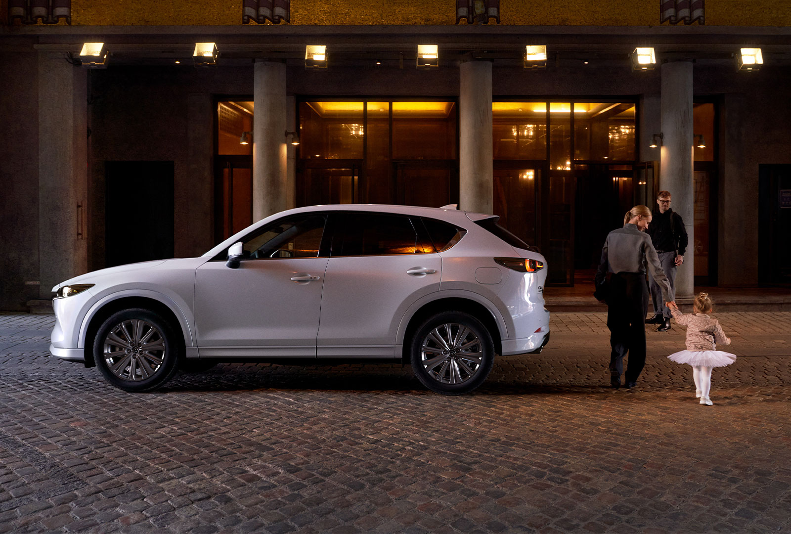 Woman escorts child in white tutu to building entrance past parked Rhodium White Metallic Mazda CX-50. 