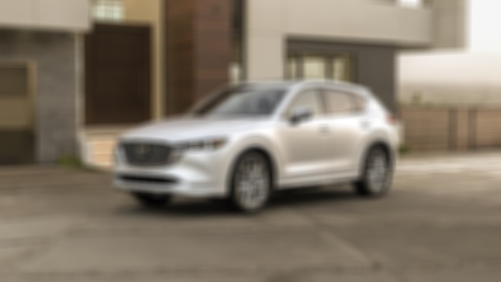 Un Mazda CX-5 blanc rhodium métallisé garé sur une allée en béton devant une maison moderne, avec en arrière-plan de vastes collines et des arbres derrière une clôture en bois. 