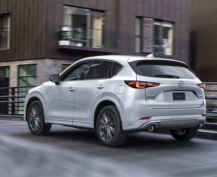 Un Mazda CX-5 blanc rhodium métallisé passe sur un viaduc avec un immeuble d’habitation surplombant la route. 