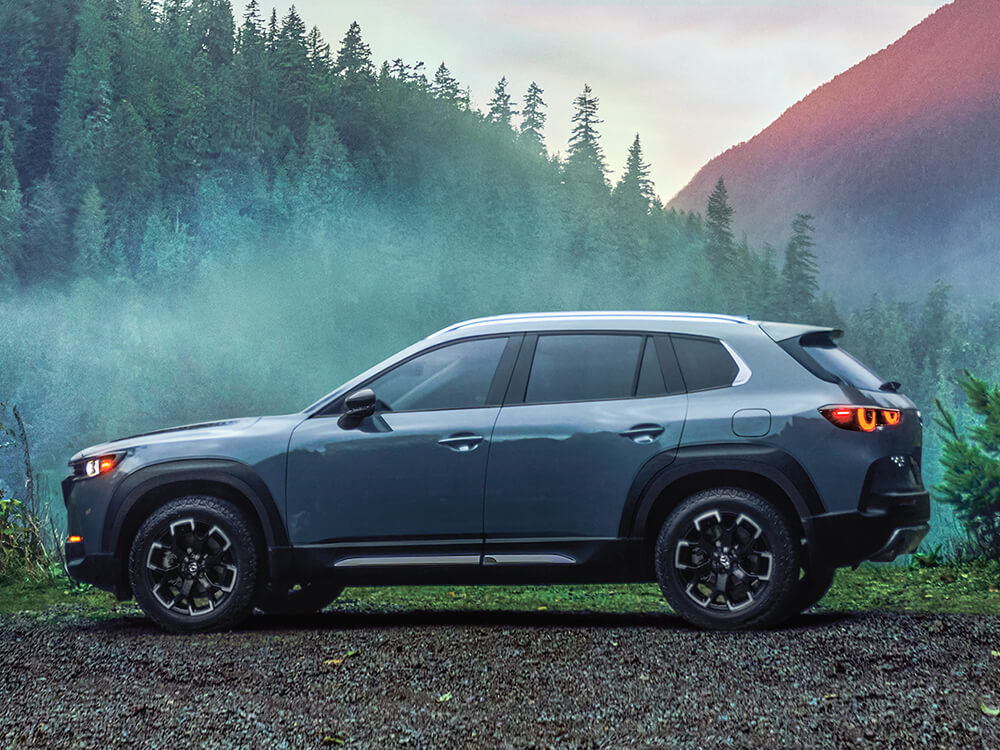 Profil d’un CX-50 Meridian garé au sommet de collines ondulantes, tourné vers le soleil qui se couche derrière des montagnes verdoyantes et brumeuses.