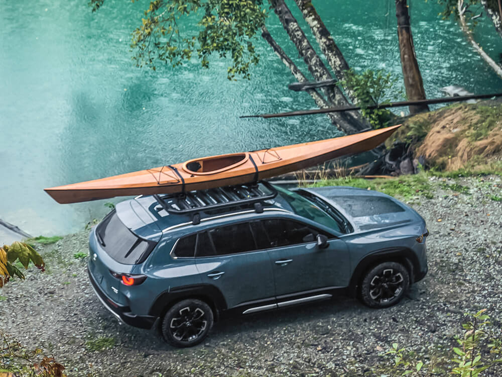 Un CX-50 Meridian garé au bord d’un lac avec les phares allumés. Un kayak est installé sur la galerie de toit.