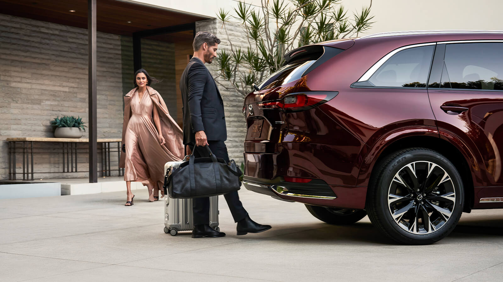 Une femme vêtue d’une robe fluide s’approche d’un homme aux mains pleines qui utilise son pied pour ouvrir le hayon mains libres d’un CX-90.  