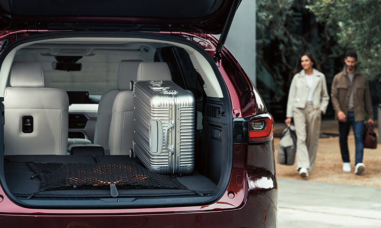 Couple approaches Artisan Red CX-90 with back lift gate open revealing a large silver suitcase.