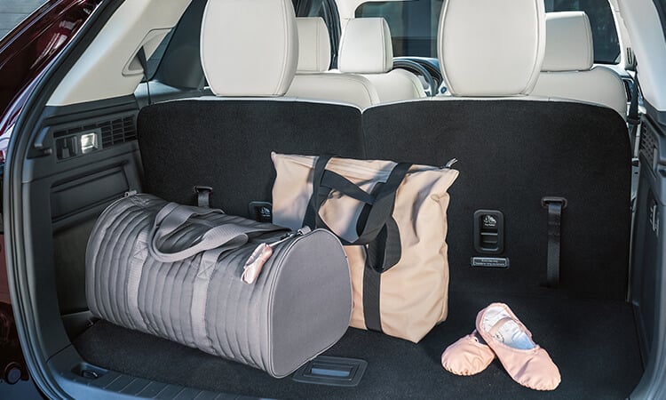 CX-90 open lift gate reveals grey duffle bag, tan laundry bag and pink ballet flats in cargo area.  