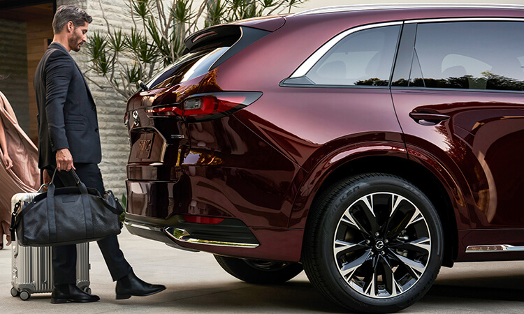 Un homme aux mains pleines utilise son pied pour activer le hayon électrique mains libres d’un CX-90 rouge artisan. 