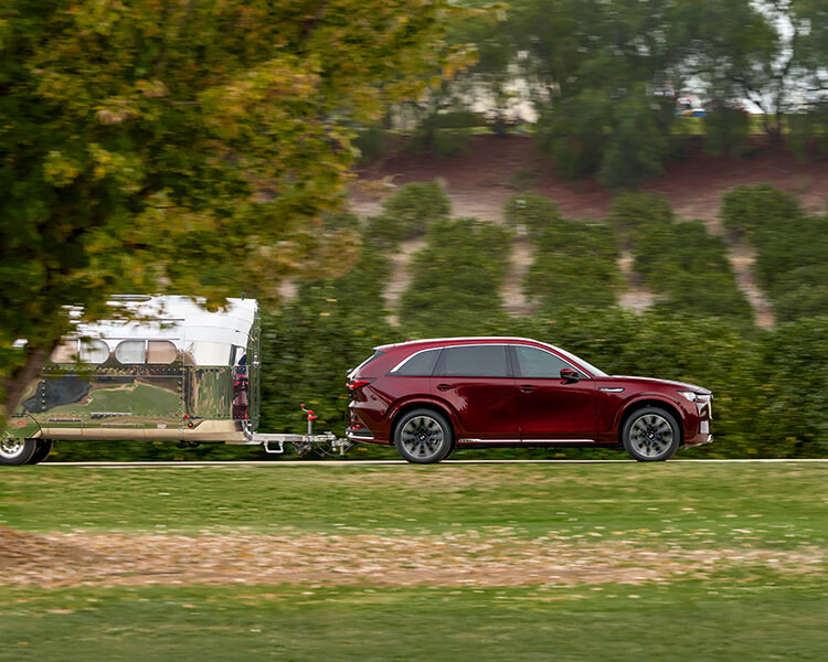 Artisan Red CX-90 PHEV tows a camper trailer along verdant backroad.