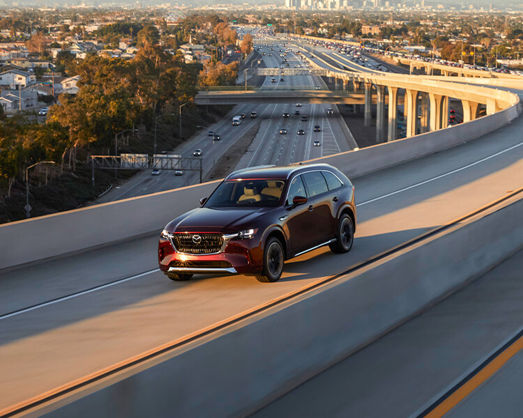 Artisan Red CX-90 PHEV drives toward sunset on concrete highway overpass. 