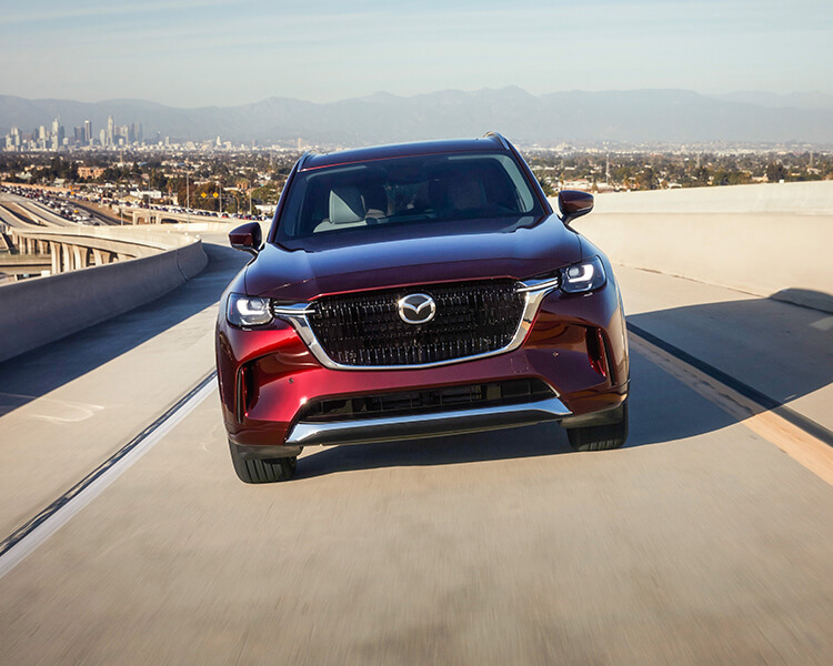  Front shot of Artisan Red CX-90 PHEV driving towards camera casting a shadow on highway overpass.