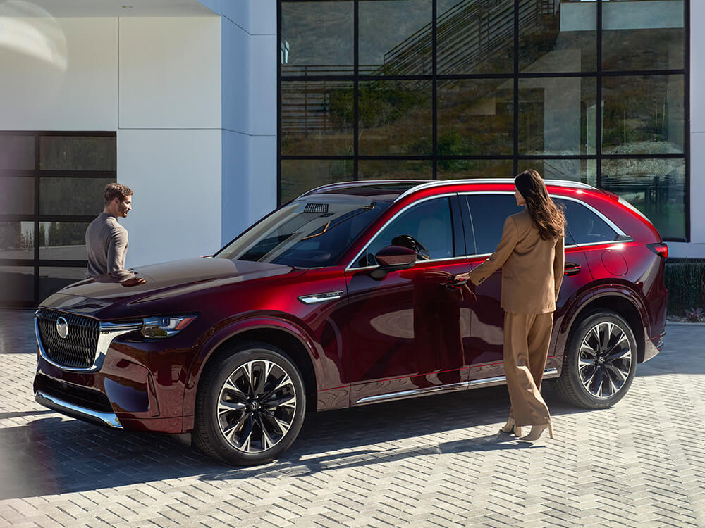 Un conducteur en costume havane ouvre la portière pour sortir d’un VÉHR CX-90 rouge artisan. 