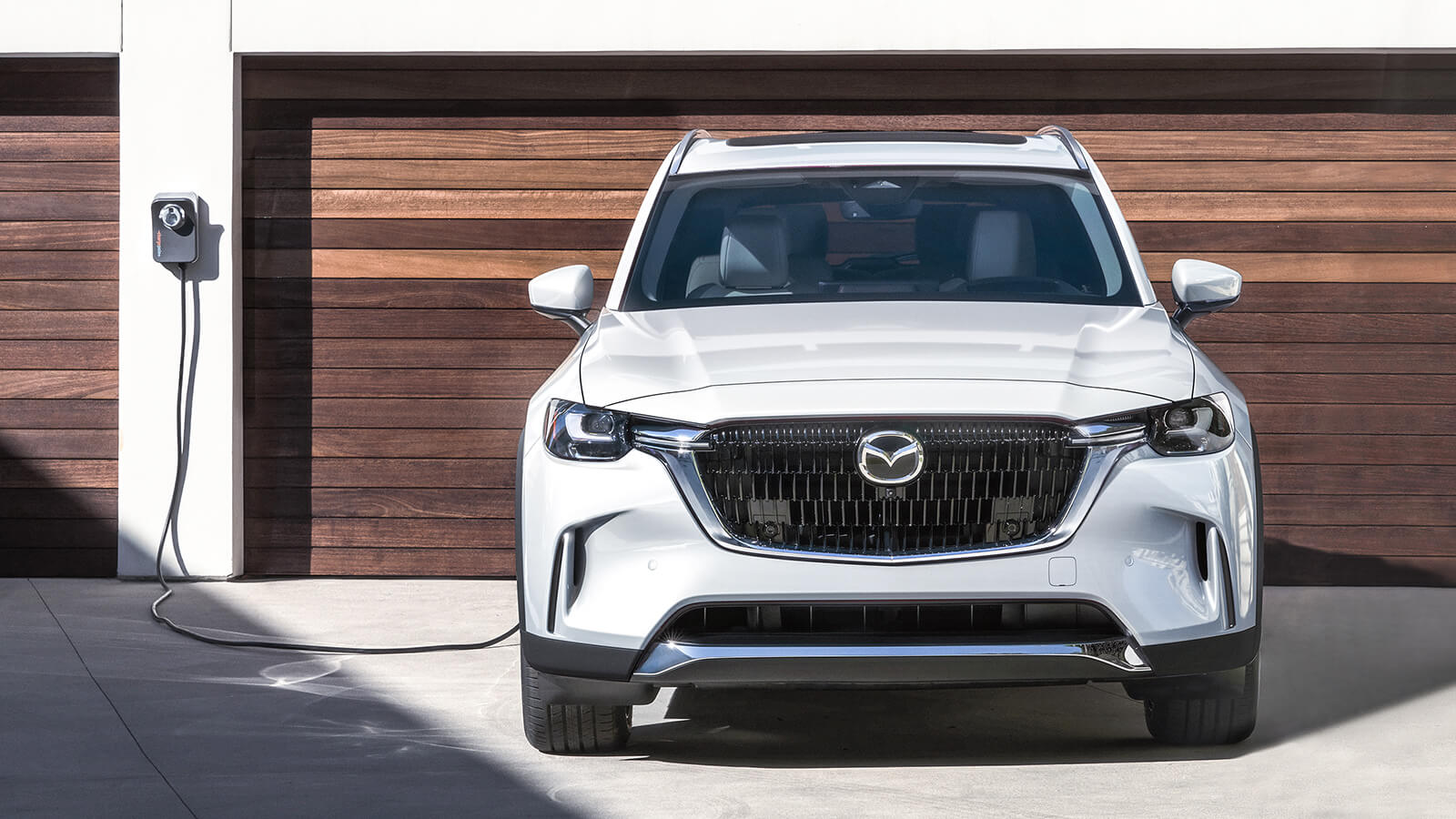 Plugged in and charging Arctic White CX-90 PHEV parked in front of double garage with wood slat garage doors.