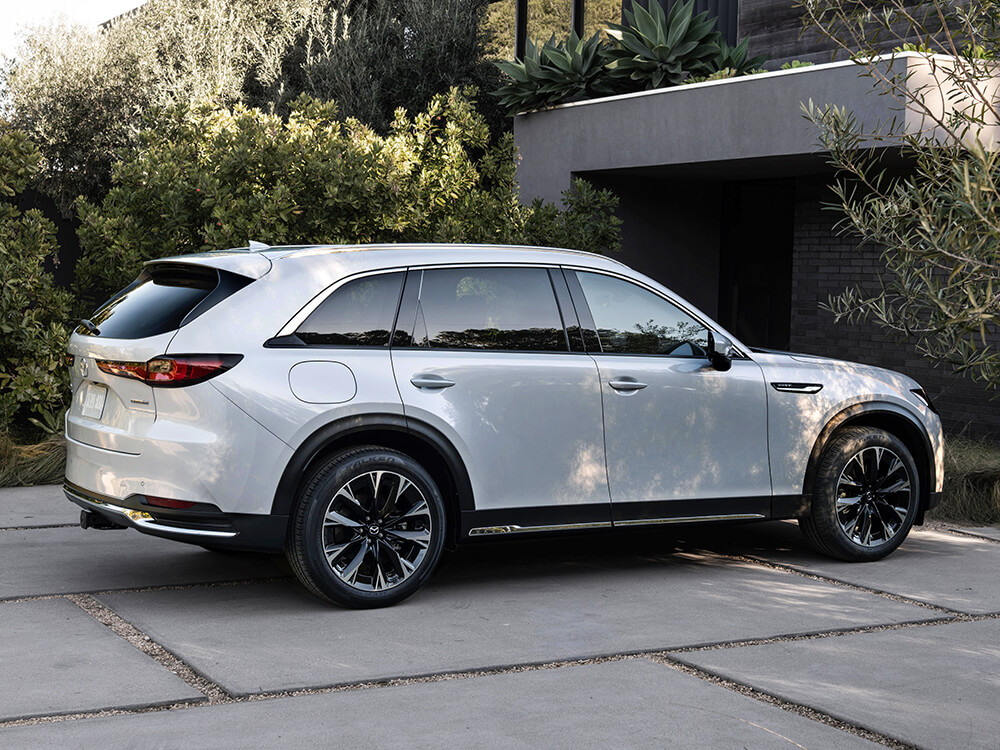 Un CX-90 blanc arctique garé dans une allée ombragée par la verdure environnante.  