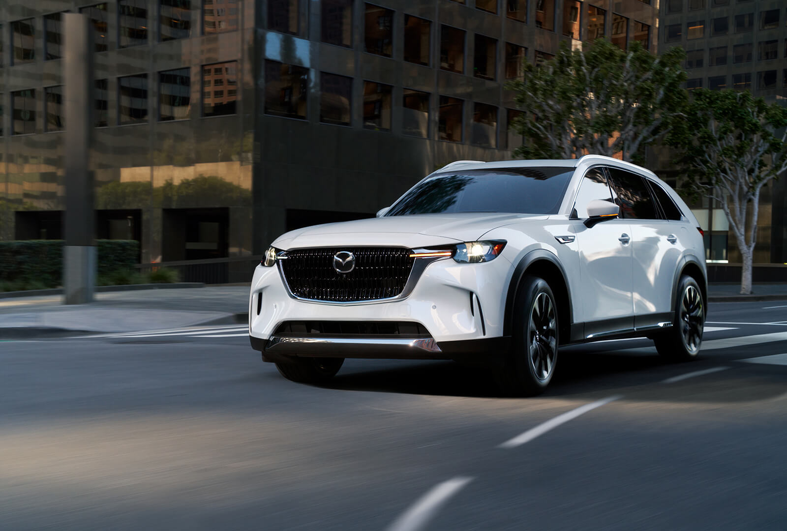 Arctic White CX-90 swiftly approaches on city street in daytime. 