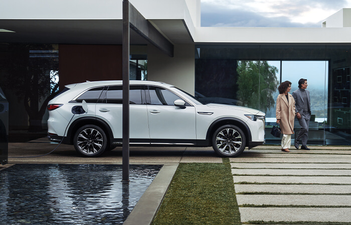 A couple walk in front of a parked and charging Arctic White CX-90. 