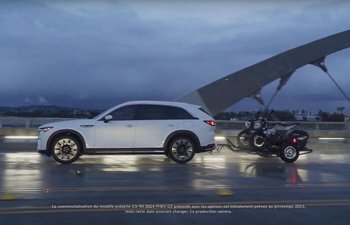 Un CX-90 PHEV roulant sur une route mouillée tout en remorquant deux motos.