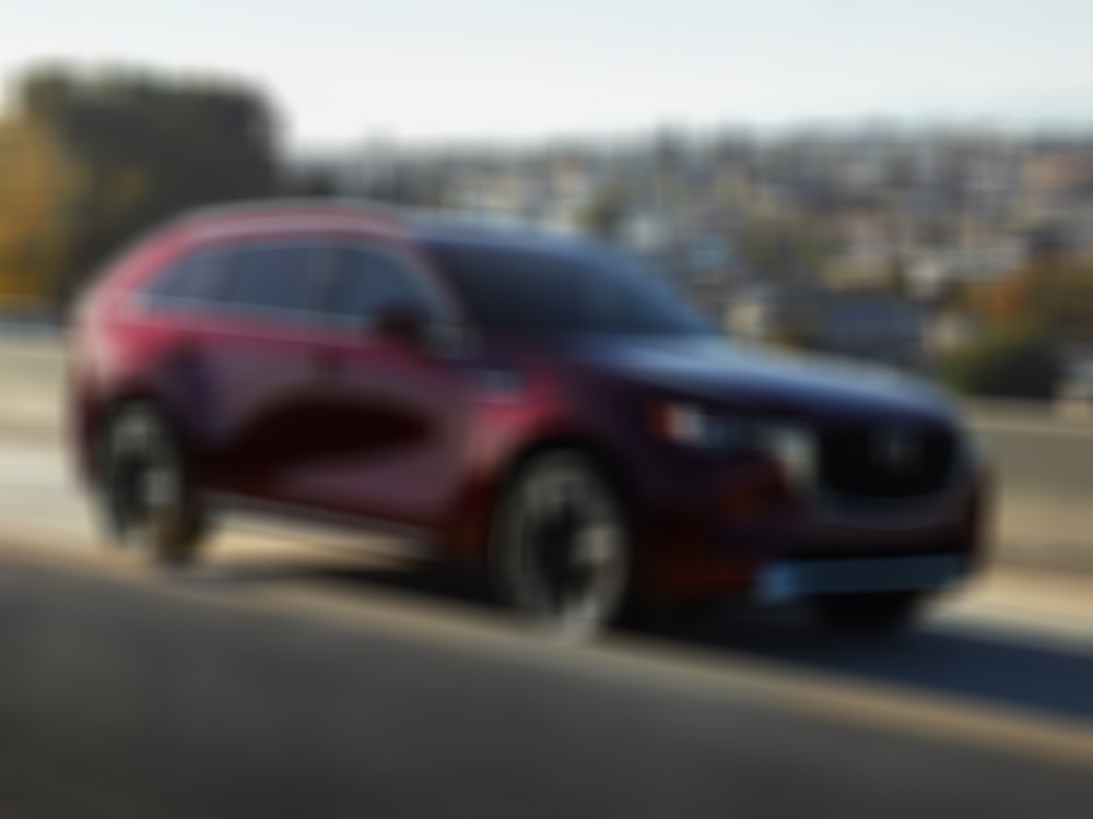 Un CX-90 rouge artisan roule sur un pont autoroutier en béton reflétant la lumière du jour.  