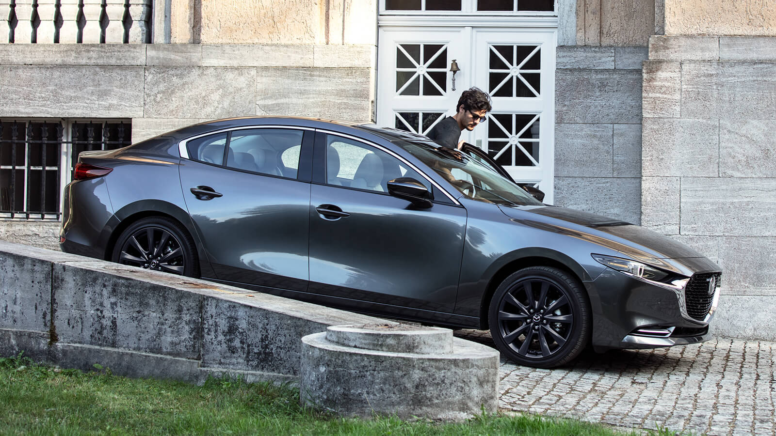 Driver stepping into a Mazda3 Sedan on cobblestone entryway in front of large limestone building. 