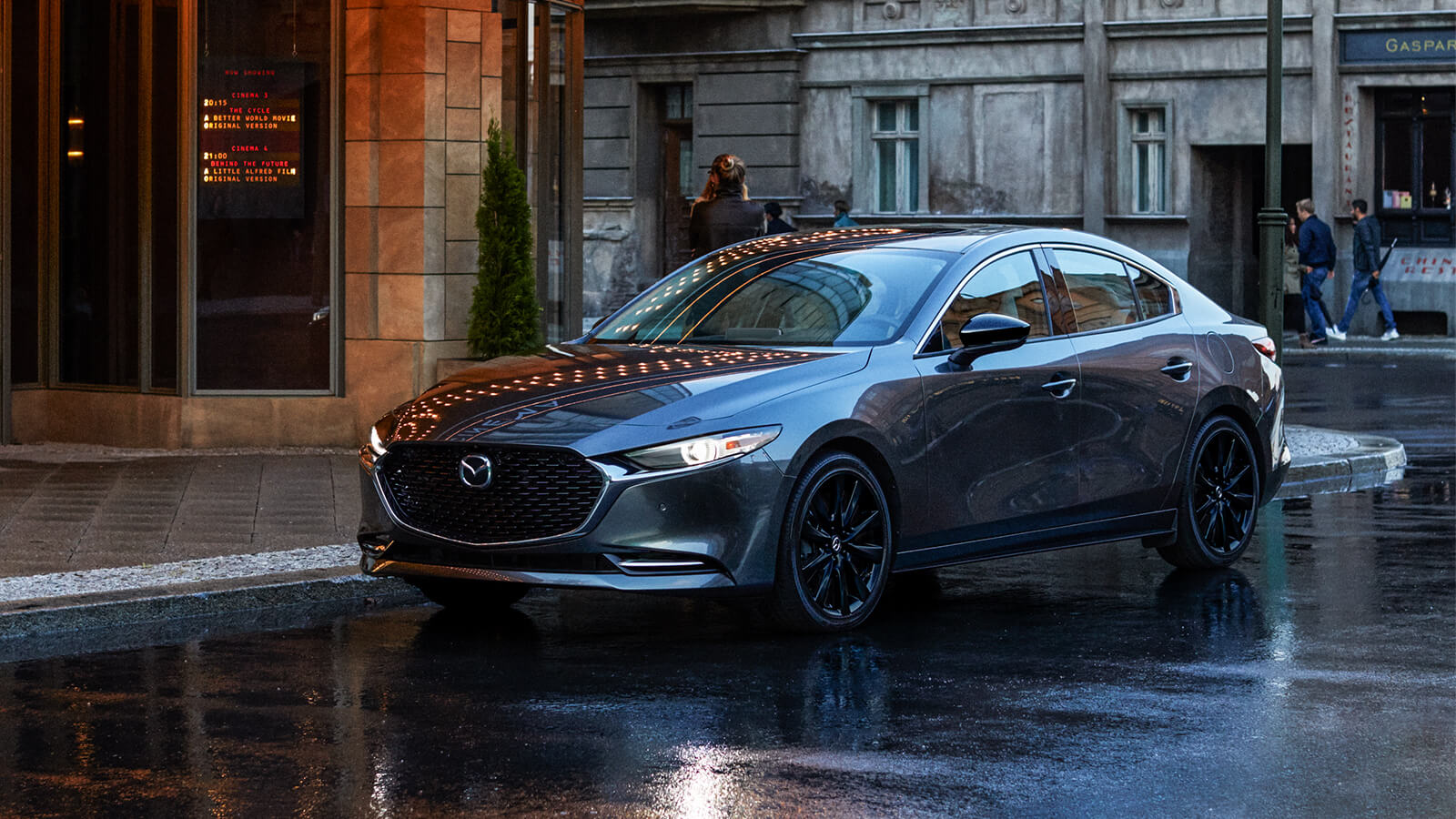 Une Mazda3 gris mécanique métallisé garée le long d’une rue urbaine, à la lumière du soleil, sur une chaussée récemment mouillée. 