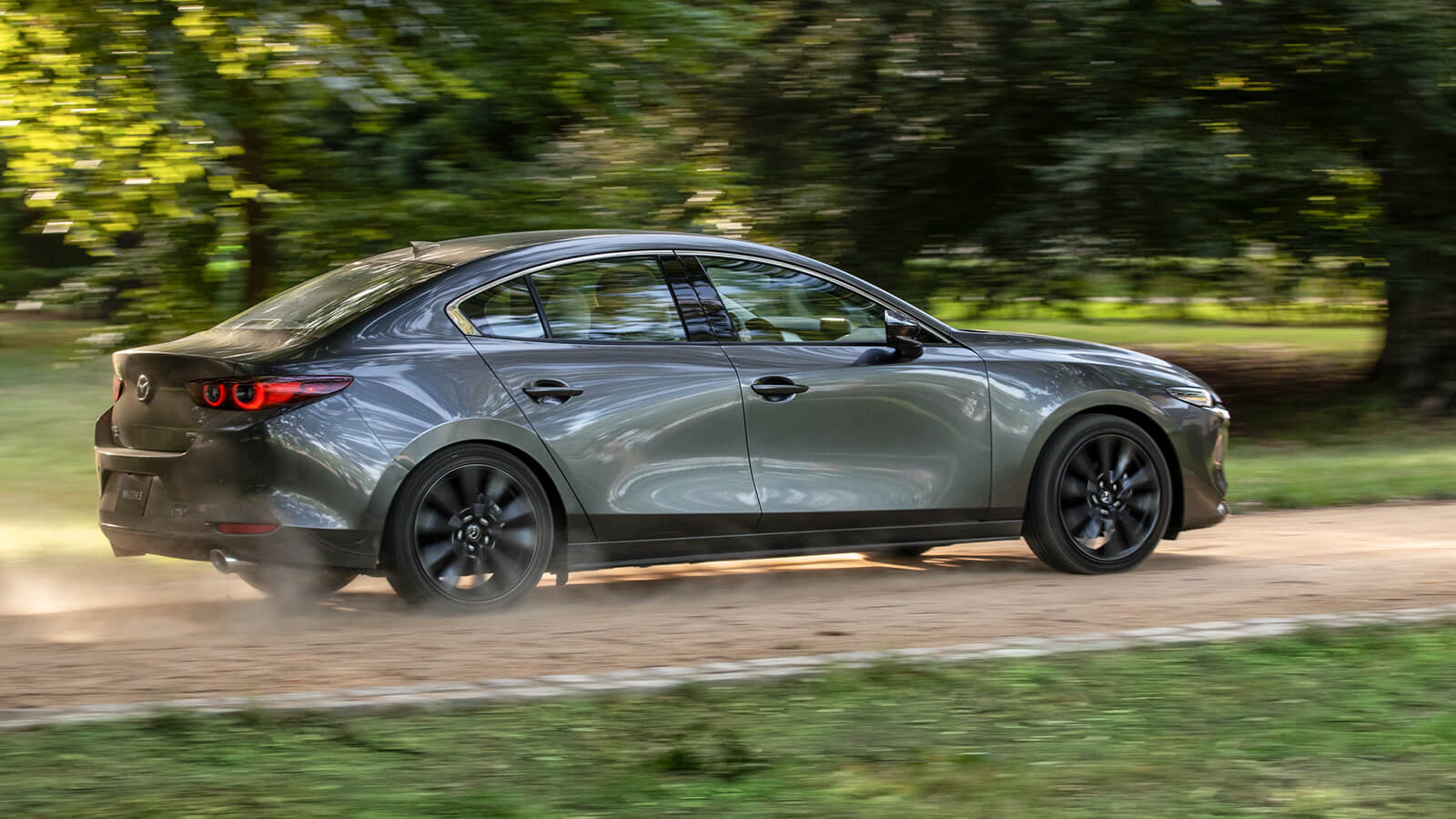 Une berline Mazda3 roule et soulève la poussière sur une route de campagne en passant devant un grand arbre en arrière-plan. 