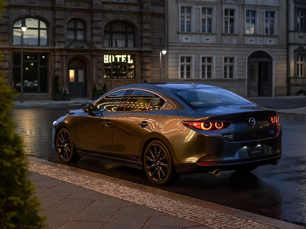 Une Mazda3 gris mécanique métallisé garée devant une salle de spectacle. La chaussée est mouillée et le véhicule reflète les lumières du chapiteau. 