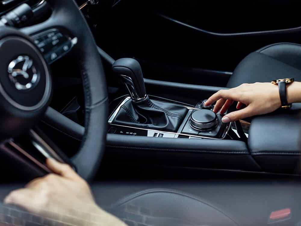 Un conducteur dans l’habitacle d’une berline Mazda3, une main tendue vers le bouton de la console centrale HMI. 