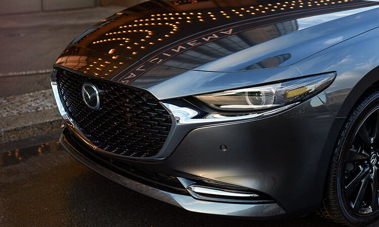 Driver’s side headlight and hood of Machine Grey Metallic Mazda3 reflecting light and skyscrapers on a city street. 