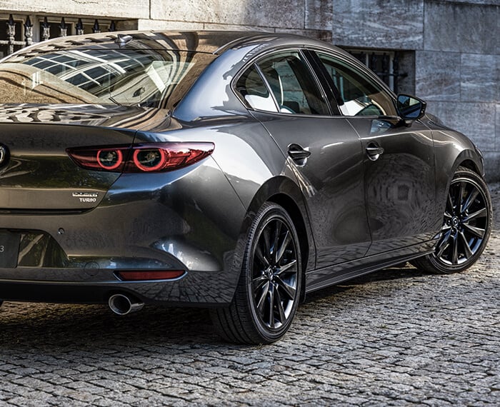 Mazda3 sedan turns its wheels to exit a cobblestone alley way in daylight.