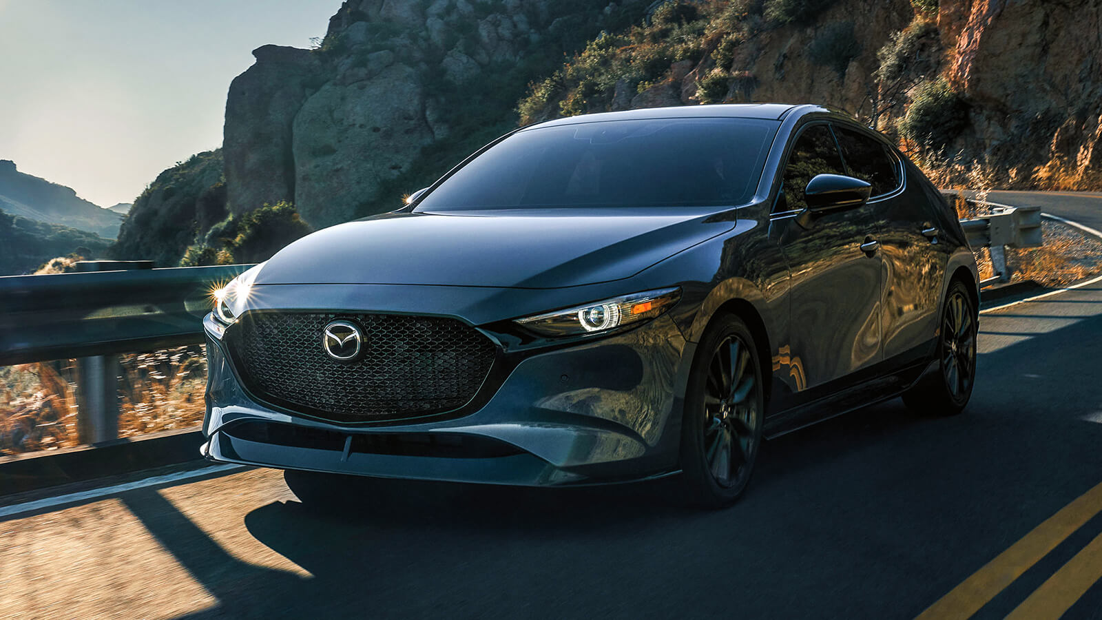 Une Mazda3 Sport gris polymétal roulant sur une autoroute de campagne, avec des collines rocheuses en arrière-plan au coucher du soleil.