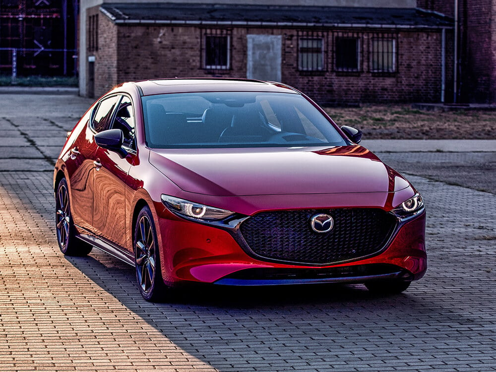 Vue avant d’une Mazda3 Sport rouge vibrant cristal métallisé garée dans une rue pavée au lever du soleil. 