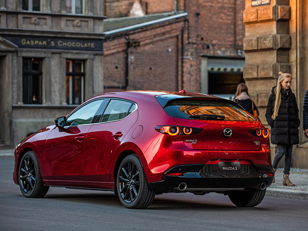 Soul Red Crystal Metallic Mazda3 Sport stopped on quaint city main street corner;, a couple passes on the sidewalk. 