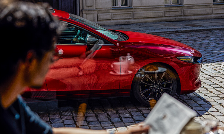 Man at cafe table on sidewalk looks at his reflection in a Soul Red Crystal Metallic Mazda3 Sport parked on cobblestone street. 