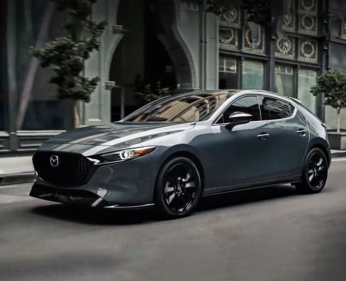 Une Mazda3 Sport descend une rue urbaine et passe devant la grande arche de l’entrée d’un hôtel.