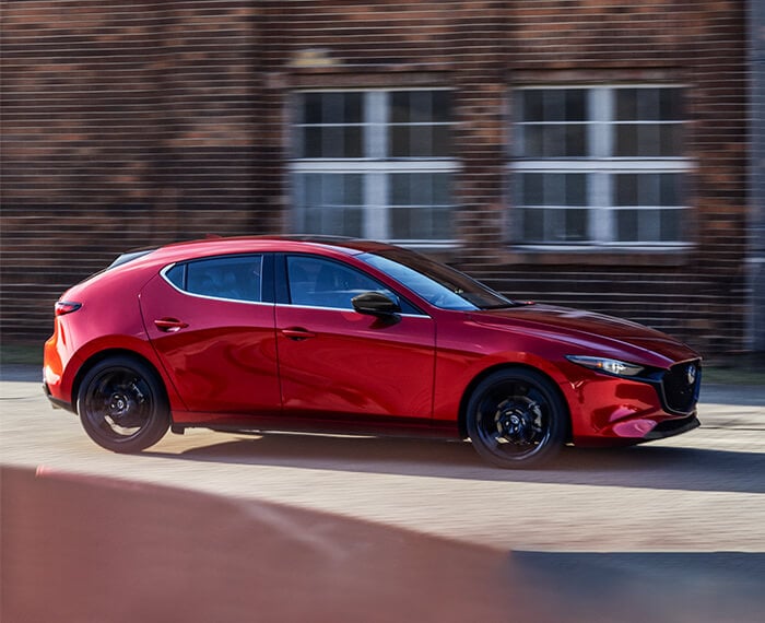 Une Mazda3 Sport à hayon de couleur rouge vibrant cristal métallisé passe devant un bâtiment en briques flou.