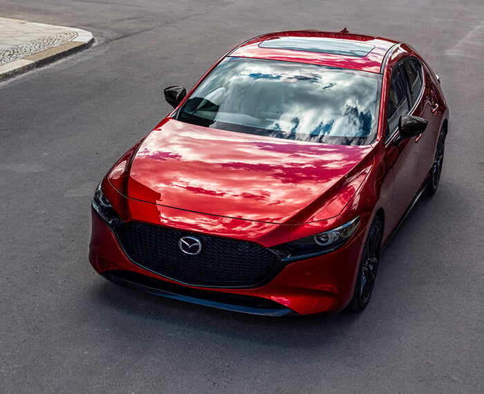 Une Mazda3 Sport rouge vibrant cristal métallisé roule sur une route pavée, reflétant le ciel bleu partiellement nuageux. 