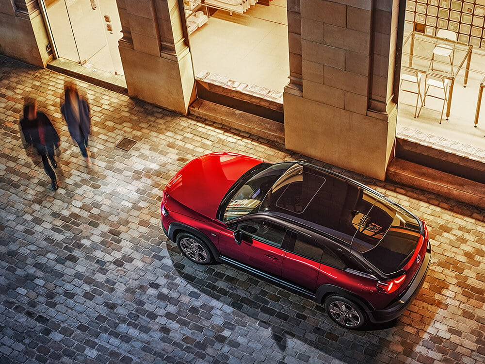 Shot of Soul Red Crystal Metallic Multi-Tone MX-30 parked in front of a store from above at night, illuminated by bright, warm lights through the store’s large windows. 