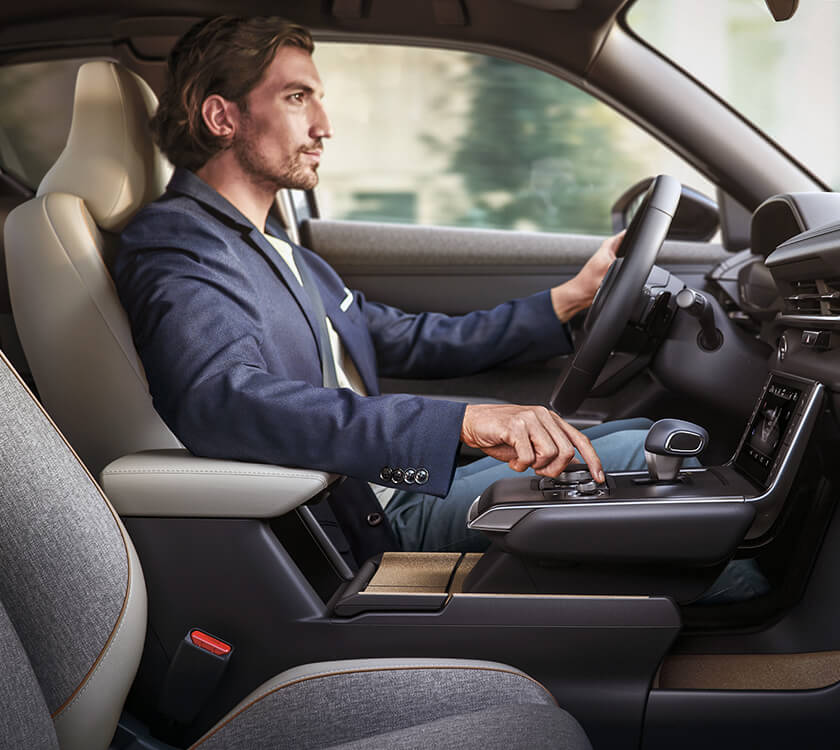 Le conducteur d’un MX-30 regarde la route devant lui, une main sur le volant et l’autre sur le bouton de commande HMI dans la console centrale.