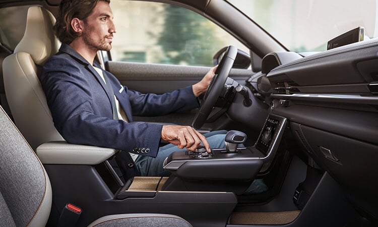 Driver in MX-30 EV looks to the road ahead with one hand on the wheel and the other controlling the HMI Command switch in the centre console.