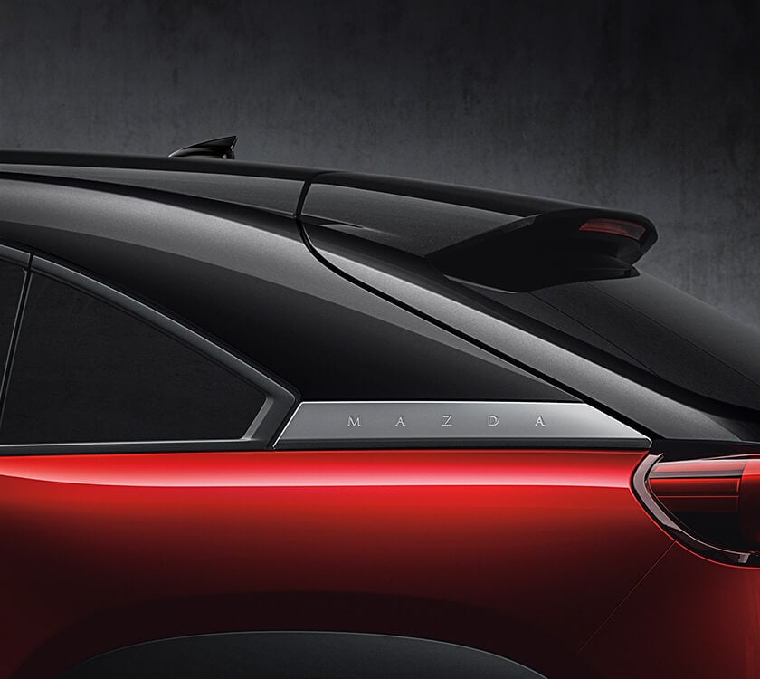 Gros plan de l’emblème « MAZDA » derrière les vitres des sièges arrière d’un MX-30 multiteinte rouge vibrant cristal métallisé.