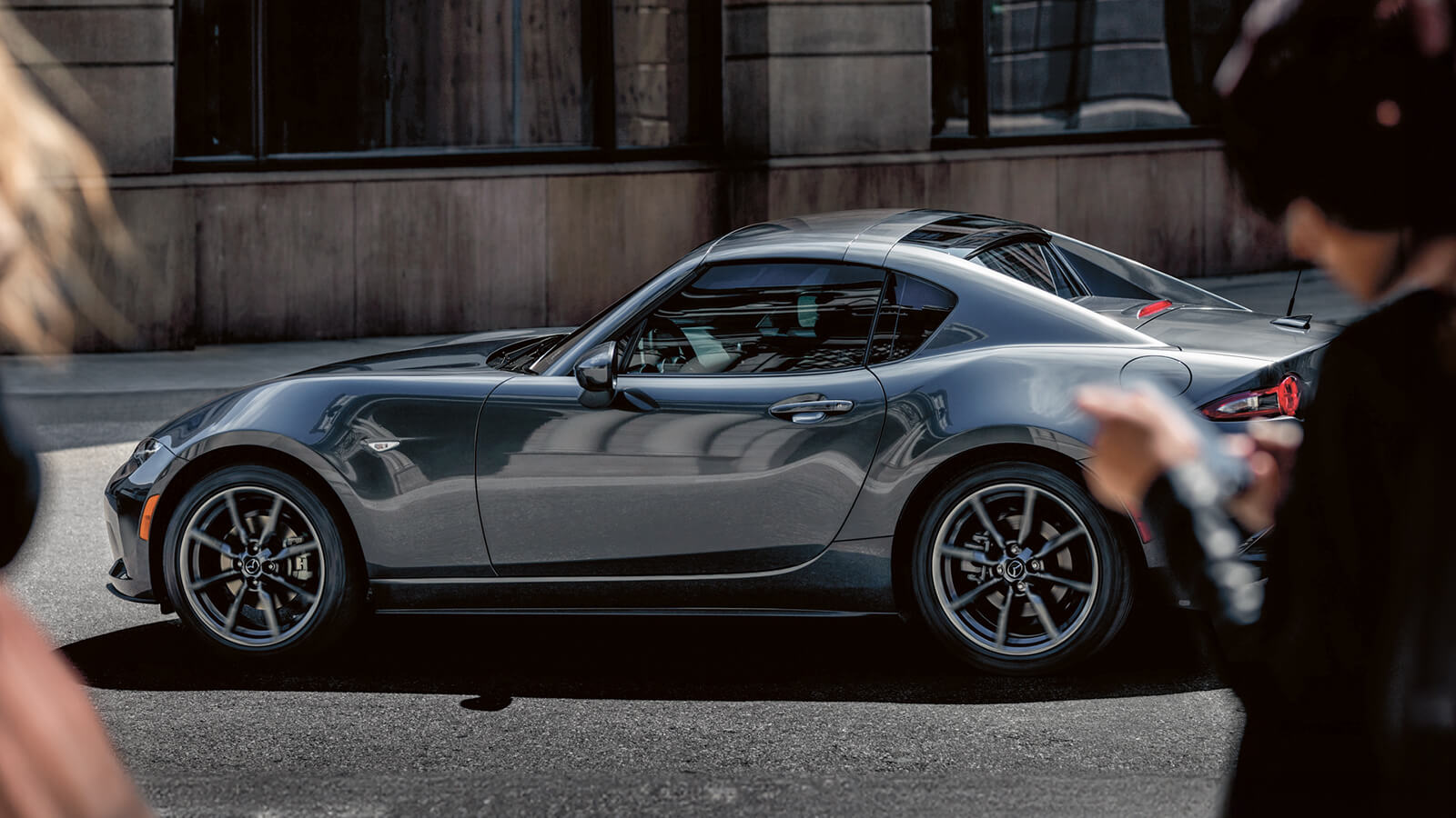 Pedestrian in foreground looks up from smart phone to watch Machine Grey Metallic MX-5 RF drive by on city street. 