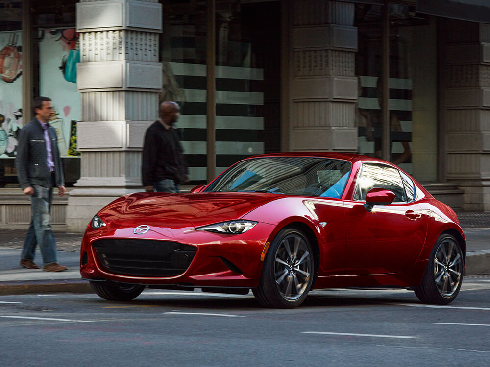 Une MX-5 RF rouge vibrant cristal métallisé avec le toit fermé s’éloigne d’un passage pour piétons; des gens attendent pour traverser en arrière-plan. 