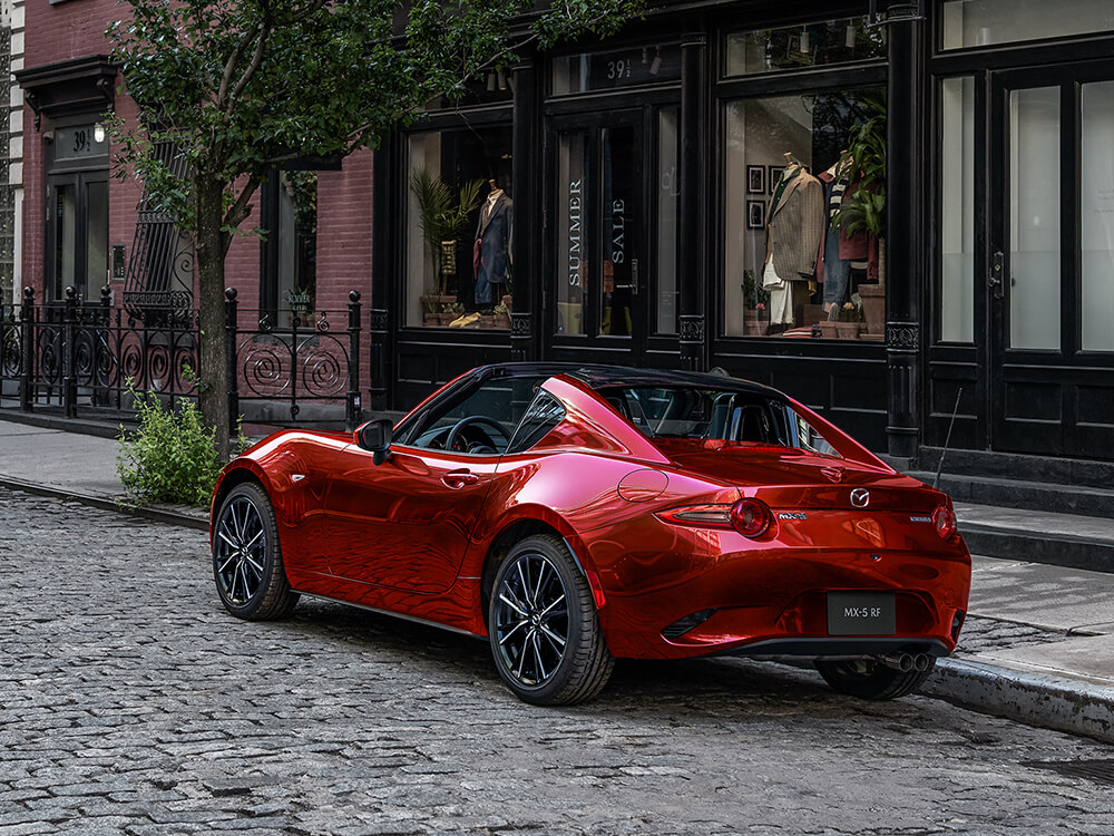 Une rutilante MX-5 RF rouge vibrant cristal métallisé, avec le toit baissé, est garée le long d’une rue pavée, devant une mercerie pour homme.