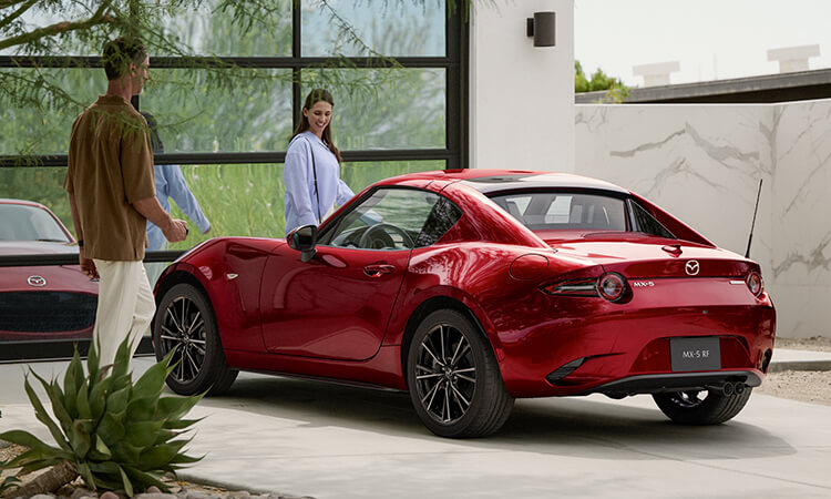 Une MX-5 RF rouge vibrant cristal métallisé avec le toit fermé garée devant une porte de garage qui s’y reflète; un couple souriant s’approche de chaque porte.