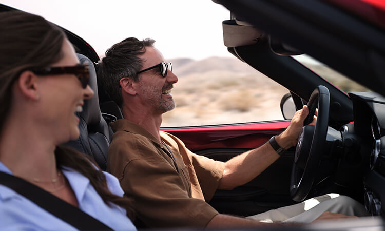 View from passenger's side inside an MX-5, passenger and driver laugh together as they drive with the top down. 