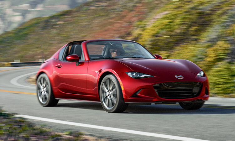 Une MX-5 RF rouge vibrant cristal métallisé avec le toit ouvert dans un virage sur une route verdoyante escarpée. 