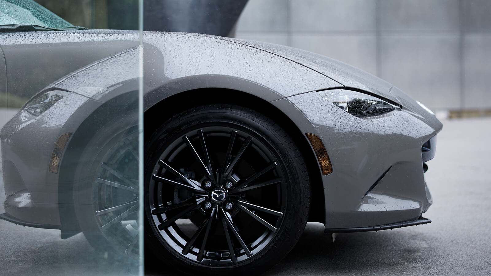 Machine Grey Metallic MX-5's front end peering out from a translucent glass panel on a rainy day.